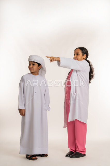 Pointing to something, specifying a specific location, ambitions and future goals of working in clinics and hospitals, portrait of an Arab Gulf Emirati girl wearing a white coat and stethoscope standing next to a boy wearing a turban and kandura with expressions of pleasure and happiness, full body image, white background