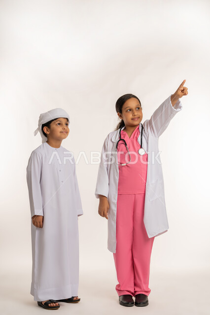 تحديد موقع معين، الاشارة الى شيء ما، الطموحات والاهداف المستقبلية بالعمل في العيادات والمشافي، بورتريه لفتاة عربية خليجية اماراتية ترتدي المعطف الأبيض والسماعات الطبية تقف بجانب ولد يرتدي العمامة والكندورة بتعابير السرور والسعادة، صورة لكامل الجسم، خلفية بيضاء