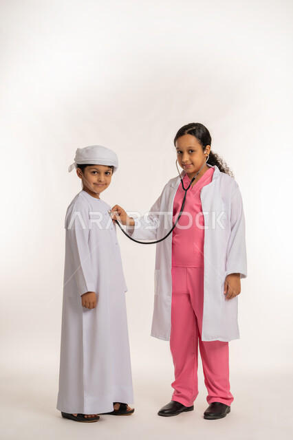 الشغف و الجد لتحقيق الأهداف، الطموحات والاهداف المستقبلية بالعمل في العيادات والمشافي، بورتريه لفتاة عربية خليجية اماراتية ترتدي المعطف الأبيض والسماعات تقوم بفحص ولد يرتدي العمامة والكندورة بتعابير السرور والسعادة، صورة لكامل الجسم، خلفية بيضاء