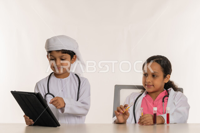 اختيار وتحديد المهنة المستقبلية منذ الصغر، استخدام جهاز تقني حديث ومتطور، اجراء تجارب وتفاعلات حيويه في المختبر، بورتريه لفتاة عربية خليجية اماراتية ترتدي المعطف الأبيض وتستخدم انابيب الاختبار، ولد اماراتي يرتدي الكندورة والعمامة يمسك بيده الجهاز اللوحي، خلفية بيضاء