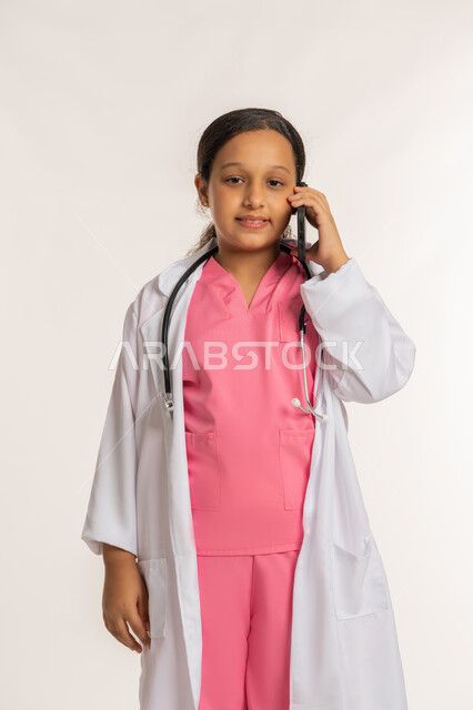 اختيار وتحديد المهنة المستقبلية منذ الصغر، بنت سعودية تقوم باجراء مكالمة هاتفية، تقديم الرعاية والخدمات الصحية، استخدام جهاز تقني حديث ومتطور، بورتريه لفتاة عربية خليجية اماراتية ترتدي المعطف الأبيض تضع السماعات الطبية  تحمل بيدها الهاتف المحمول، خلفية بيضاء