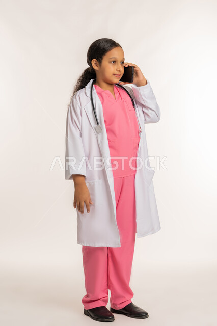 استخدام جهاز تقني حديث ومتطور، تقديم الرعاية والخدمات الصحية، بورتريه لفتاة عربية خليجية اماراتية ترتدي المعطف الأبيض تضع السماعات الطبية  تحمل بيدها الهاتف المحمول، اختيار وتحديد المهنة المستقبلية منذ الصغر، بنت سعودية تقوم باجراء مكالمة هاتفية، خلفية بيضاء
