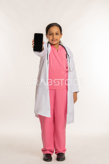 تحديد واختيار المهنة المستقبلية منذ الصغر، تقديم الرعاية والخدمات الصحية، بورتريه لفتاة عربية خليجية اماراتية ترتدي المعطف الأبيض تضع السماعات الطبية تحمل بيدها الهاتف المحمول، بنت سعودية تقوم بعرض شاشة سوداء فارغة، صورة لكامل الجسم، خلفية بيضاء
