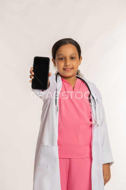 تحديد واختيار المهنة المستقبلية منذ الصغر، تقديم الرعاية والخدمات الصحية، بورتريه لفتاة عربية خليجية اماراتية ترتدي المعطف الأبيض تضع السماعات الطبية تحمل بيدها الهاتف المحمول، بنت سعودية تقوم بعرض شاشة سوداء فارغة، صورة لكامل الجسم، خلفية بيضاء