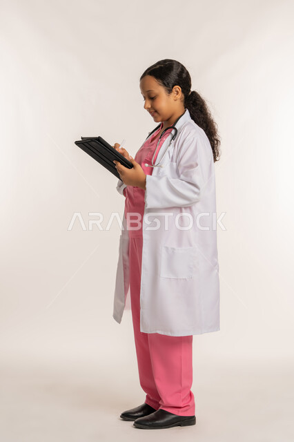 استخدام قلم التابليت لتدوين الملاحظات، الطموح والهدف المستقبلي بالعمل في العيادات والمشافي، كتابة نتائج الفحوصات، بورتريه لفتاة عربية خليجية اماراتية ترتدي معطف أبيض تحمل بيدها الجهاز اللوحي، بنت سعودية  تقوم بإعداد التقارير الطبيه، صورة لكامل الجسم، خلفية بيضاء