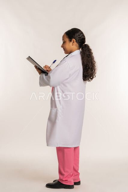 Ambition and future goal of working in clinics and hospitals, determining future career, side portrait of an Arab Gulf Emirati girl wearing a white coat recording test results in the medical record, Saudi girl preparing medical reports, full body, white background