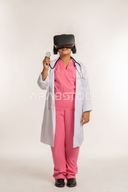 تحديد المهنة المستقبلية منذ الصغر، مفهوم الطب والرعاية الصحية، الاندماج مع العالم الخيالي، بورتريه لفتاة عربية خليجية اماراتية ترتدي المعطف الأبيض تضع نظارة الفي ار تقوم بالتفاعل مع الواقع الافتراضي، صبية سعودية تمسك بيدها السماعة، الجسم بالطول الكامل، خلفية بيضاء