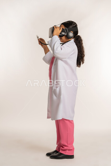 مفهوم الطب والرعاية الصحية، الاندماج مع العالم الخيالي، تحديد المهنة المستقبلية منذ الصغر، بورتريه من الجانب لفتاة عربية خليجية اماراتية ترتدي المعطف الأبيض تضع نظارة الفي ار تقوم بالتفاعل مع الواقع الافتراضي، صبية سعودية تمسك بيدها السماعة، خلفية بيضاء