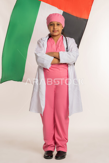 ذكرى اليوم الوطني الإماراتي 2 ديسمبر, الاحتفال بالأعياد والمناسبات الوطنية, علم الامارات العربية المتحدة،  بورتريه لبنت عربية خليجية اماراتية ترتدي المعطف الأبيض تضع السماعات الطبية تقف بأيدي متقاطعة بايماءات الثقة بالنفس, صورة لكامل الجسم، خلفية بيضاء