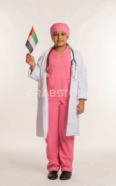 دور الاطباء في المجتمع، ذكرى اليوم الوطني الإماراتي 2 ديسمبر, الاحتفال بالأعياد والمناسبات الوطنية، بورتريه لبنت عربية خليجية اماراتية ترتدي المعطف الأبيض تضع السماعات الطبية تحمل بيدها علم الامارات العربية المتحدة, صورة لكامل الجسم، خلفية بيضاء