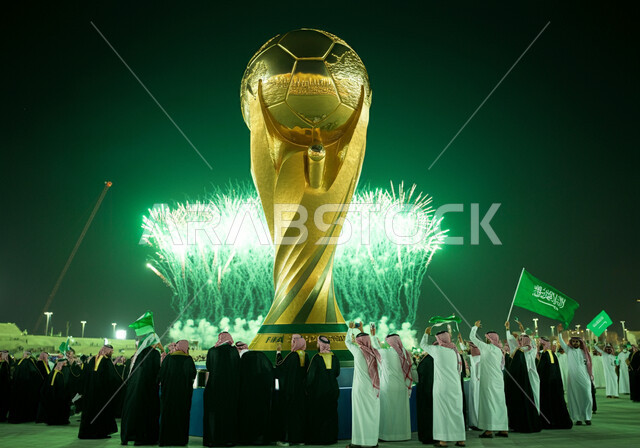 الاحتفالات بفوز السعوديه باستضافة كاس العالم 2034، مجسم كأس العالم الذهبي لرياضة كرة القدم، الاهتمام بالرياضة والانشطة الترفيهية، مجموعة من الرجال العرب الخليجيون السعوديون يرتدون الثوب التقليدي يطلقون المفرقعات والالعاب النارية في الليل