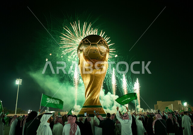 مجموعة من الرجال العرب الخليجيون السعوديون يرتدون الثوب التقليدي يطلقون المفرقعات والالعاب النارية في الليل، الاهتمام بالرياضة والانشطة الترفيهية، الاحتفالات بفوز السعوديه باستضافة كاس العالم 2034، مجسم كأس العالم الذهبي لرياضة كرة القدم