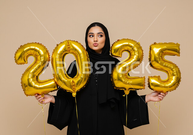 إيماءات السعادة والسرور, البهجة بقدوم العام الجديد, التفاؤل والترحيب بالسنة الجديدة, الاحتفال بعيد رأس السنة الميلادية 2025, بورتريه لامرأة عربية خليجية سعودية مبتسمة محجبة ترتدي العباءة السوداء تحمل بيدها بالونات ذهبية كبيرة, خلفية بيج