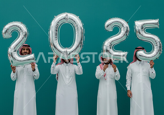 التفاؤل والترحيب بالسنة الجديدة, البهجة بقدوم العام الجديد, الاحتفال بعيد رأس السنة الميلادية 2025, إيماءات السعادة والسرور, بورتريه لرجال عرب خليجيين سعوديين يرتدون الثوب التقليدي والشماغ يحملون بأيديهم بالونات فضية كبيرة, خلفية زرقاء