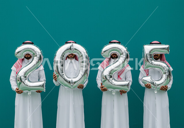 البهجة بقدوم العام الجديد, الاحتفال بعيد رأس السنة الميلادية 2025, إيماءات السعادة والسرور, التفاؤل والترحيب بالسنة الجديدة, بورتريه لرجال عرب خليجيين سعوديين يرتدون الثوب التقليدي والشماغ يحملون بأيديهم بالونات فضية كبيرة, خلفية زرقاء
