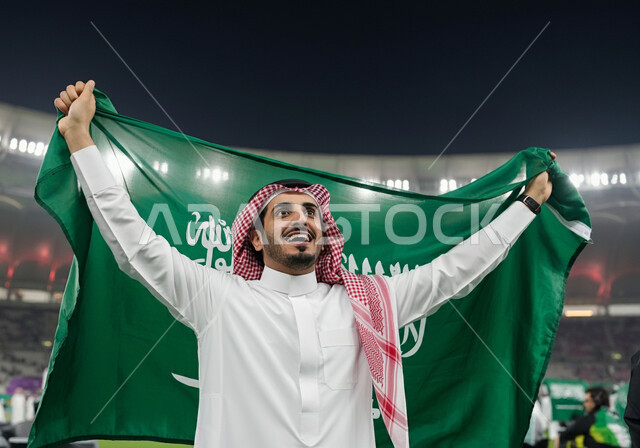 استضافة الأولومبياد الرياضية، بطولة كأس العالم 2034 ، مونديال المملكة العربية السعودية فيفا, تشجيع الفريق السعودي, صورة مقربة لرجل عربي خليجي سعودي يرتدي الثوب التقليدي والشماغ يحمل على ظهره علم السعودية بتعابير الحماس