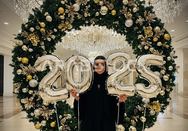 النظر إلى الكاميرا بإيماءات السعادة والسرور, الاحتفال بعيد رأس السنة الميلادية 2025, مفهوم التفاؤل والترحيب بالسنة الجديدة, امرأة عربية خليجية سعودية مبتسمة محجبة ترتدي العباءة السوداء تقف أمام إطار مزين وتحمل بيديها بالونات فضية كبيرة