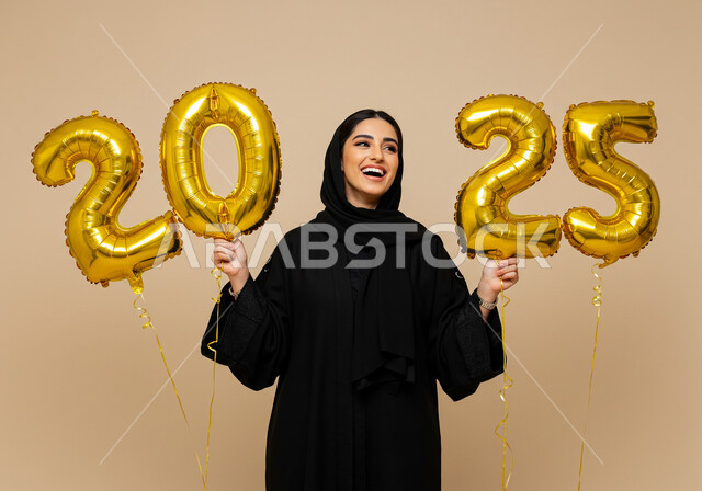 الاحتفال بعيد رأس السنة الميلادية 2025, البهجة بقدوم العام الجديد, إيماءات السعادة والسرور, التفاؤل والترحيب بالسنة الجديدة, بورتريه لامرأة عربية خليجية سعودية مبتسمة محجبة ترتدي العباءة السوداء تحمل بيدها بالونات ذهبية كبيرة, خلفية بيج