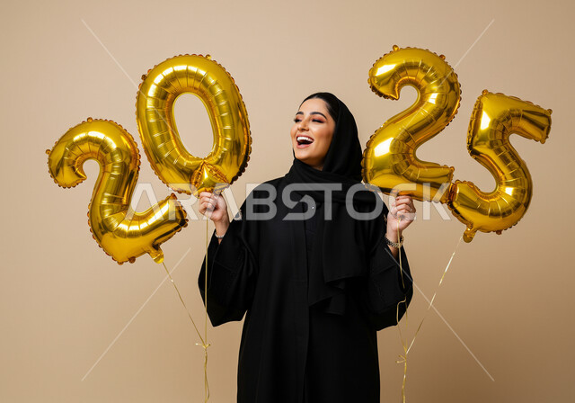 الاستعداد لاستقبال العام الجديد, الاحتفال بعيد رأس السنة الميلادية 2025, تعابير الفرح والسعادة والسرور, البهجة والتفاؤل والترحيب بالسنة الجديدة, بورتريه لامرأة عربية خليجية سعودية مبتسمة محجبة ترتدي العباءة السوداء تحمل بيدها بالونات ذهبية كبيرة, خلفية بيج