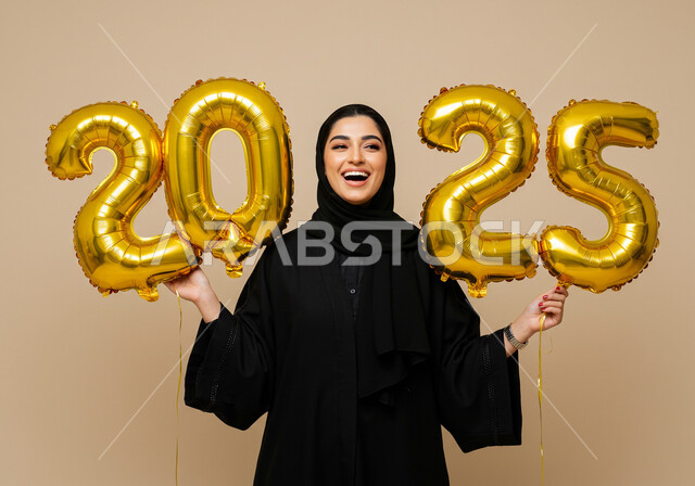 البهجة والتفاؤل والترحيب بالسنة الجديدة, تعابير الفرح والسعادة والسرور, الاحتفال بعيد رأس السنة الميلادية 2025, بورتريه لامرأة عربية خليجية سعودية مبتسمة محجبة ترتدي العباءة السوداء تحمل بيدها بالونات ذهبية كبيرة, الاستعداد لاستقبال العام الجديد, خلفية بيج