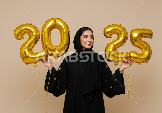 الاستعداد لاستقبال العام الجديد, الاحتفال بعيد رأس السنة الميلادية 2025, تعابير الفرح والسعادة والسرور, البهجة والتفاؤل والترحيب بالسنة الجديدة, بورتريه لامرأة عربية خليجية سعودية مبتسمة محجبة ترتدي العباءة السوداء تحمل بيدها بالونات ذهبية كبيرة, خلفية بيج