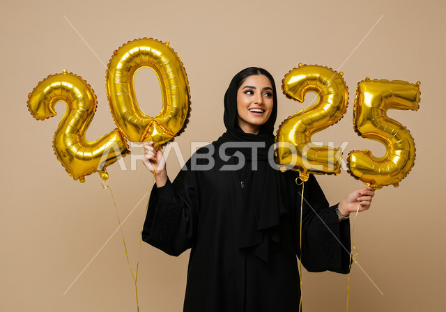 البهجة والتفاؤل والترحيب بالسنة الجديدة, تعابير الفرح والسعادة والسرور, الاحتفال بعيد رأس السنة الميلادية 2025, بورتريه لامرأة عربية خليجية سعودية مبتسمة محجبة ترتدي العباءة السوداء تحمل بيدها بالونات ذهبية كبيرة, الاستعداد لاستقبال العام الجديد, خلفية بيج