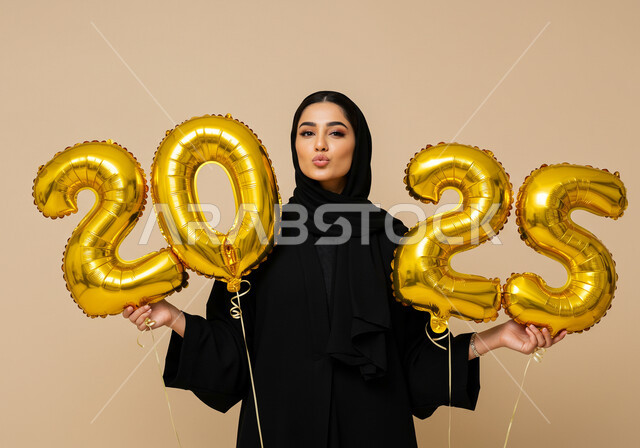 تعابير الفرح والسعادة والسرور, البهجة والتفاؤل والترحيب بالسنة الجديدة, الاحتفال بعيد رأس السنة الميلادية 2025, بورتريه لامرأة عربية خليجية سعودية محجبة ترتدي العباءة السوداء تحمل بيدها بالونات ذهبية كبيرة, الاستعداد لاستقبال العام الجديد, خلفية بيج