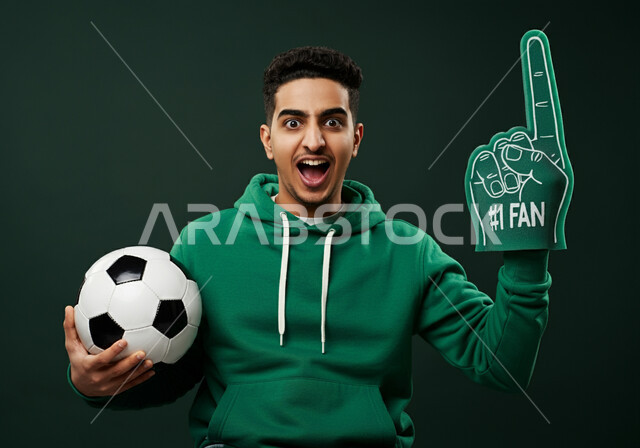 الشغف بتشجيع الفريق المفضل، النظر الى الكاميرا بإيماءات السرور والحماس لتحقيق الفوز و الانتصار، بورتريه مقرب لشاب عربي خليجي سعودي يرتدي قفاز تشجيعي اخضر عليه رقم واحد يحمل بيده كرة القدم، متابعه المبارايات السعودية، خلفية خضراء