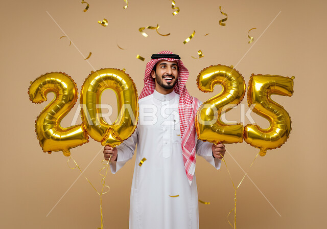 التفاؤل والترحيب بالسنة الجديدة, إيماءات السعادة والسرور, الاستعداد لاستقبال العام الجديد, بورتريه لرجل عربي خليجي سعودي مبتسم يرتدي الثوب التقليدي والشماغ يحمل بيديه بالونات ذهبية كبيرة, الاحتفال بعيد رأس السنة الميلادية 2025, خلفية بيج