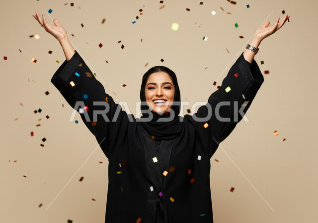 قصاصات صغيرة ملونة, أوراق زينة ذهبية, النظر إلى الكاميرا بإيماءات السعادة والسرور,, الاحتفال بالأعياد والمناسبات السعيدة, بورتريه مقرب لامرأة عربية خليجية سعودية مبتسمة محجبة ترتدي العباءة السوداء ترفع يديها للأعلى بتعابير البهجة والفرح, خلفية بيج