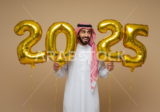 إيماءات السعادة والسرور, التفاؤل والترحيب بالسنة الجديدة, الاستعداد لاستقبال العام الجديد, بورتريه لرجل عربي خليجي سعودي مبتسم يرتدي الثوب التقليدي والشماغ يحمل بيديه بالونات ذهبية كبيرة, الاحتفال بعيد رأس السنة الميلادية 2025, خلفية بيج