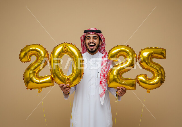 الاستعداد لاستقبال العام الجديد, إيماءات السعادة والسرور, التفاؤل والترحيب بالسنة الجديدة, بورتريه لرجل عربي خليجي سعودي مبتسم يرتدي الثوب التقليدي والشماغ يحمل بيديه بالونات ذهبية كبيرة, الاحتفال بعيد رأس السنة الميلادية 2025, خلفية بيج