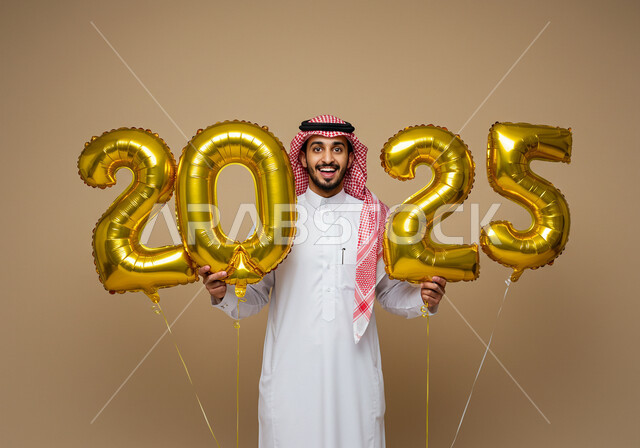 الاحتفال بعيد رأس السنة الميلادية 2025, الاستعداد لاستقبال العام الجديد, إيماءات السعادة والسرور, التفاؤل والترحيب بالسنة الجديدة, بورتريه لرجل عربي خليجي سعودي مبتسم يرتدي الثوب التقليدي والشماغ يحمل بيديه بالونات ذهبية كبيرة, خلفية بيج