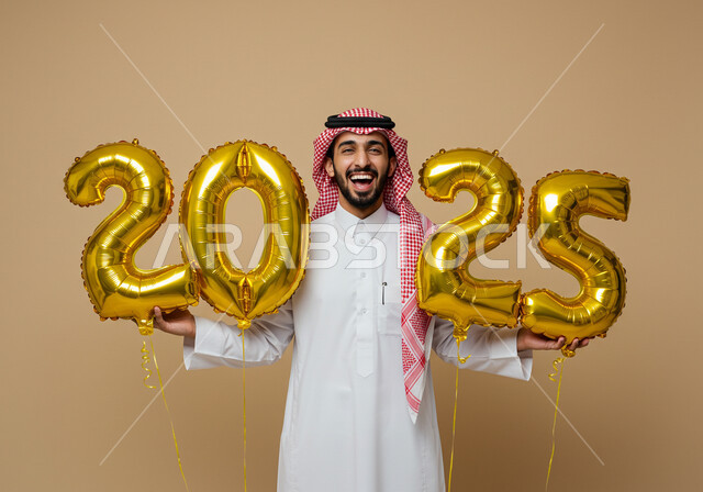إيماءات السعادة والسرور, التفاؤل والترحيب بالسنة الجديدة, الاستعداد لاستقبال العام الجديد, بورتريه لرجل عربي خليجي سعودي مبتسم يرتدي الثوب التقليدي والشماغ يحمل بيديه بالونات ذهبية كبيرة, الاحتفال بعيد رأس السنة الميلادية 2025, خلفية بيج