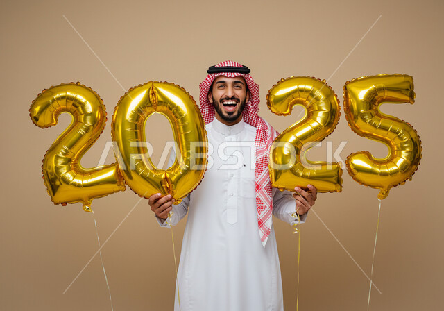 الاستعداد لاستقبال العام الجديد, إيماءات السعادة والسرور, التفاؤل والترحيب بالسنة الجديدة, بورتريه لرجل عربي خليجي سعودي مبتسم يرتدي الثوب التقليدي والشماغ يحمل بيديه بالونات ذهبية كبيرة, الاحتفال بعيد رأس السنة الميلادية 2025, خلفية بيج