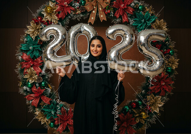 الاحتفال بعيد رأس السنة الميلادية 2025, النظر إلى الكاميرا بإيماءات السعادة والسرور, مفهوم التفاؤل والترحيب بالسنة الجديدة, امرأة عربية خليجية سعودية مبتسمة محجبة ترتدي العباءة السوداء تقف أمام إطار مزين وتحمل بيديها بالونات فضية كبيرة
