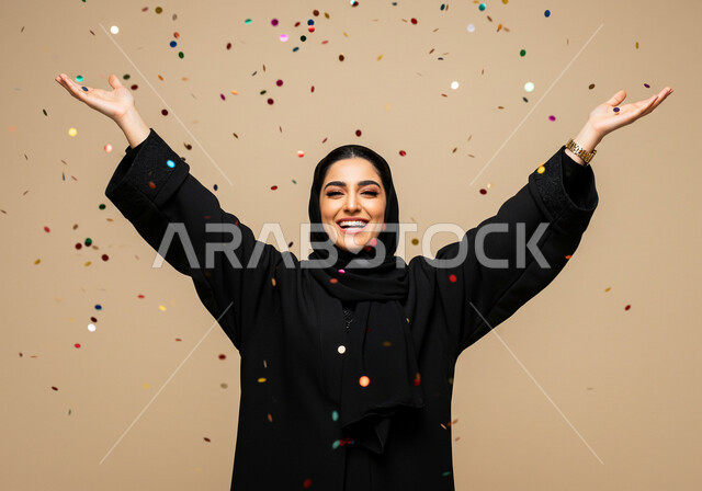 النظر إلى الكاميرا بإيماءات السعادة والسرور, أوراق زينة ذهبية, قصاصات صغيرة ملونة, الاحتفال بالأعياد والمناسبات السعيدة, بورتريه مقرب لامرأة عربية خليجية سعودية مبتسمة محجبة ترتدي العباءة السوداء ترفع يديها للأعلى بتعابير البهجة والفرح, خلفية بيج