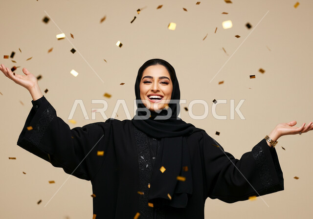 الاحتفال بالأعياد والمناسبات السعيدة, النظر إلى الكاميرا بإيماءات السعادة والسرور, أوراق زينة ذهبية, قصاصات صغيرة ملونة, بورتريه مقرب لامرأة عربية خليجية سعودية مبتسمة محجبة ترتدي العباءة السوداء ترفع يديها للأعلى بتعابير البهجة والفرح, خلفية بيج