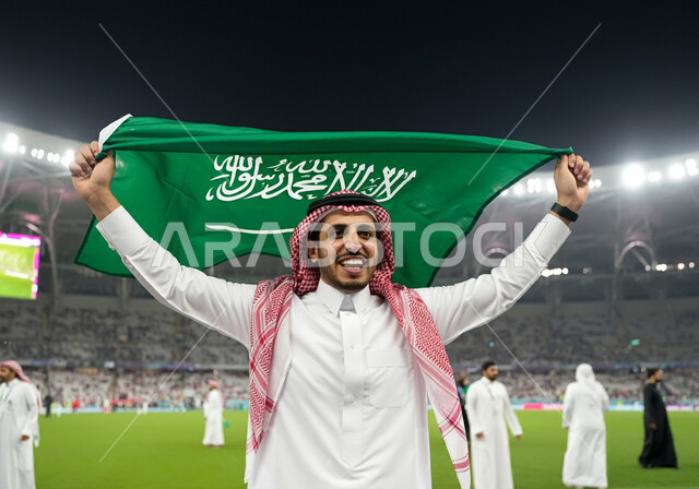 اليوم الوطني السعودي 23 سبتمبر, ذكرى يوم العلم 11 مارس، رجل عربي خليجي سعودي يرتدي الثوب التقليدي والشماغ يحمل علم المملكة ويقف في الملعب، تشجيع المنتخب السعودي،  الاهتمام بالرياضة والانشطة الترفيهية، ايماءات الحماس والسرور، الاحتفال بالأعياد والمناسبات الوطنية