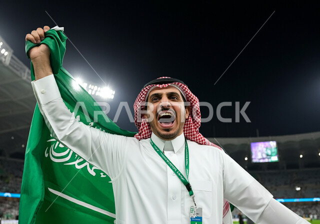 تشجيع المنتخب السعودي، ايماءات الحماس والسرور، الاحتفال بالأعياد والمناسبات الوطنية، اليوم الوطني السعودي 23 سبتمبر, ذكرى يوم العلم 11 مارس، رجل عربي خليجي سعودي يرتدي الثوب التقليدي والشماغ يحمل علم المملكة ويقف في الملعب، الاهتمام بالرياضة والانشطة الترفيهية