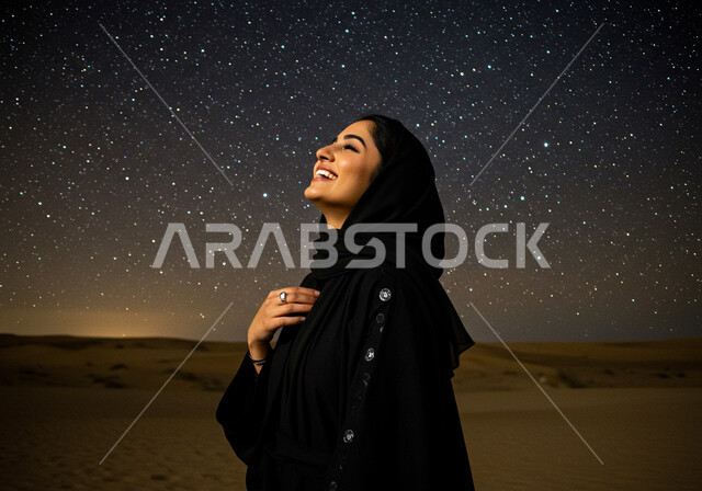 الاستمتاع بالجو الليلي الهادئ, مغامرة برية في صحاري السعودية, الرمال الذهبية الناعمة, رحلة سياحية ممتعة وسط الصحراء, صورة مقربة لامرأة عربية خليجية سعودية محجبة ترتدي العباءة السوداء تنظر إلى السماء بإيماءات السعادة والسرور, مجرة درب التبانة في الليل