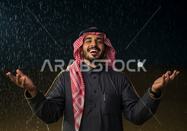 الاستمتاع بالوقوف تحت الأمطار, الجو الشتوي الماطر في صحاري المملكة العربية السعودية, الرمال الذهبية الناعمة في الصحراء, صورة مقربة لرجل عربي خليجي سعودي يرتدي الثوب التقليدي والشماغ يقف تحت المطر الغزير ويرفع يديه للأعلى بتعابير السعادة والسرور