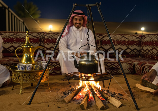 الجو الليلي الهادئ, رجل عربي خليجي سعودي يرتدي الثوب التقليدي والشماغ يجلس أمام شعلة الحطب، مخيم سياحي في منطقة صحراوية، كشتة و جلسة بر شبابية, التشكيلات والكثبان الرملية, الرمال الذهبية الناعمة، رحلة تخييم سياحية ممتعة وسط الصحراء