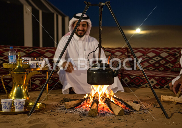 ابريق شاي على الحطب، رحلة تخييم سياحية ممتعة وسط الصحراء، الجو الليلي الهادئ, رجل عربي خليجي سعودي يرتدي الثوب التقليدي والشماغ يجلس أمام النيران المشتعلة، مخيم سياحي في منطقة صحراوية، كشتة و جلسة بر شبابية, التشكيلات والكثبان الرملية, الرمال الذهبية الناعمة