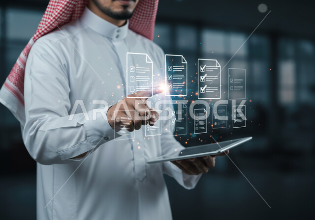 دمج التقنيات الحديثة في العمل، استخدام جهاز تقني حديث ومتطور، صورة مقربة لرجل عربي خليجي سعودي يرتدي الثوب التقليدي والشماغ يمسك بيده الايباد يقوم بالنقر على أوراق مهام هولوجرامية، رؤية مستقبلية سعودية 2030 