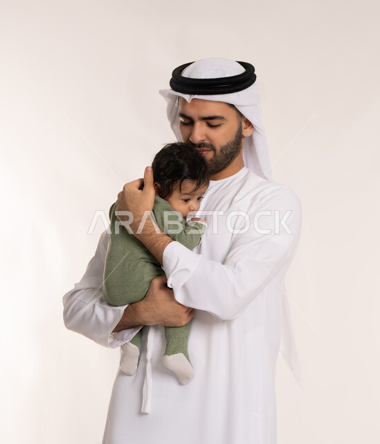 إيماءات الحب والحنان على الصغار, تحمل المسؤولية والعناية بالأطفال، بورتريه مقرب لأب عربي خليجي إماراتي يرتدي الكندورة والغترة يقوم باحتضان ابنه الرضيع بيديه, قضاء وقت ممتع مع الطفل, ملاعبة الأبناء بعطف واهتمام، خلفية بيضاء
