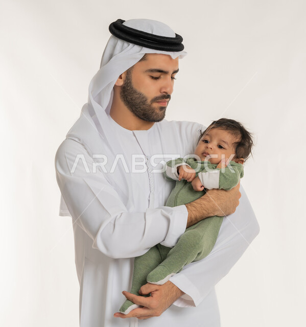 ملاعبة الأبناء بعطف واهتمام، قضاء وقت ممتع مع الطفل, بورتريه مقرب لأب عربي خليجي إماراتي يرتدي الكندورة والغترة يقوم باحتضان ابنه الرضيع بيديه, إيماءات الحب والحنان على الصغار, تحمل المسؤولية والعناية بالأطفال، خلفية بيضاء