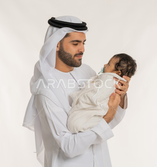 إيماءات الحب والحنان على الصغار, تحمل المسؤولية والعناية بالأطفال، بورتريه مقرب لأب عربي خليجي إماراتي يرتدي الكندورة والغترة يقوم باحتضان ابنه الرضيع بيديه, قضاء وقت ممتع مع الطفل, ملاعبة الأبناء بعطف واهتمام، خلفية بيضاء