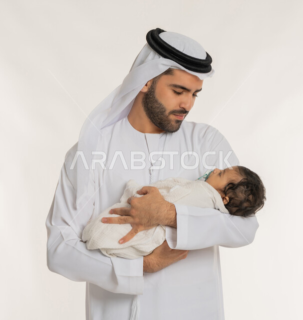 تحمل المسؤولية والعناية بالأطفال، بورتريه مقرب لأب عربي خليجي إماراتي يرتدي الكندورة والغترة يقوم باحتضان ابنه الرضيع بيديه, قضاء وقت ممتع مع الطفل, إيماءات الحب والحنان على الصغار, ملاعبة الأبناء بعطف واهتمام، خلفية بيضاء