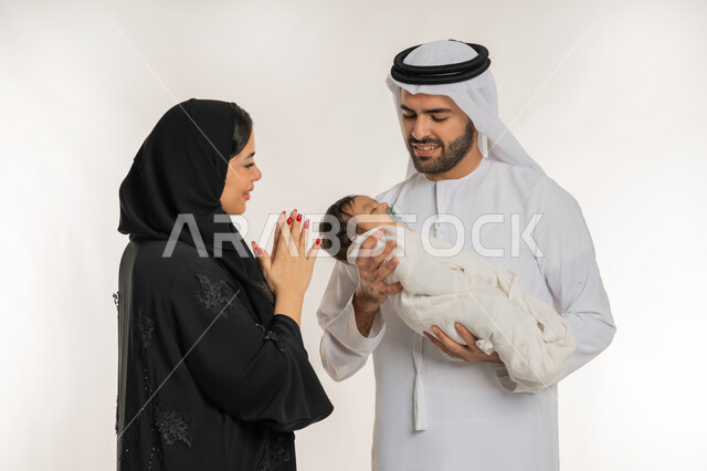 أب وأم يحتضنان طفلهما الرضيع بحب وحنان، قضاء وقت عائلي ممتع، بورتريه مقرب لعائلة عربية خليجية إماراتية يقضون وقتهم سويا, مفهوم الأسرة السعيدة المترابطة ,الرعاية والاهتمام بالأطفال، الأمان و العطف على الأبناء، أجواء أسرية مليئة بالمودة, خلفية بيضاء